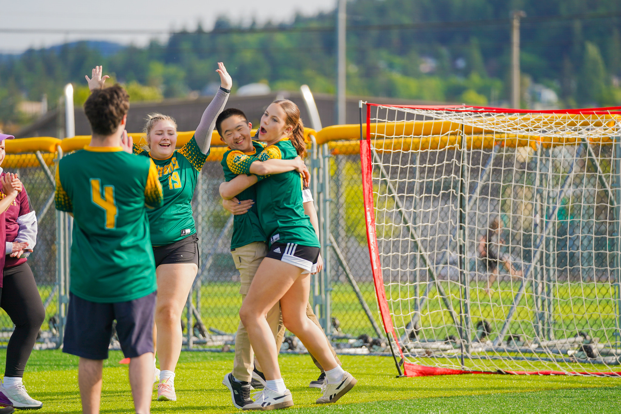 team celebrating a goal on the soccer field, smiling and hugging near the goal net during a sunny day in Bellingham, Washington